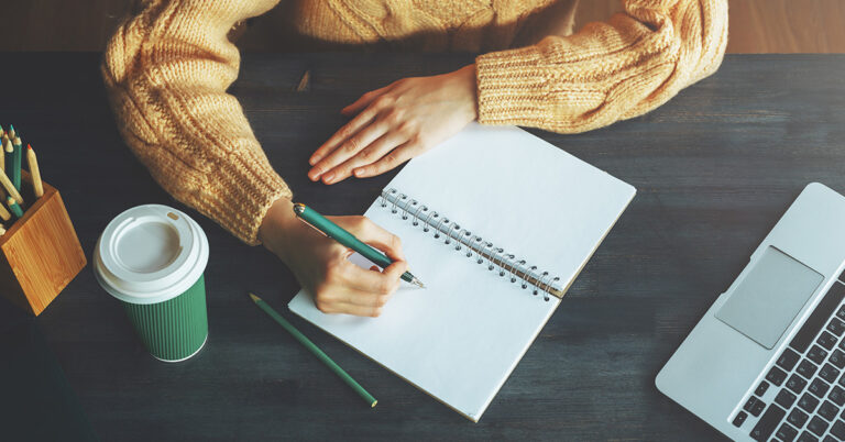 A person writing in a notebook and a coffee next to them